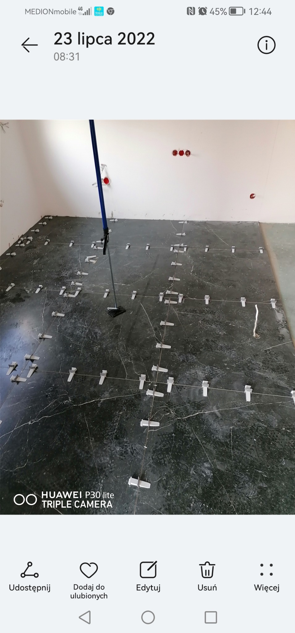 Układanie ciemnych płytek podłogowych z użyciem systemu poziomowania, widoczne sznurki i klipsy dystansowe, w tle surowe ściany z wystającymi przewodami elektrycznymi.