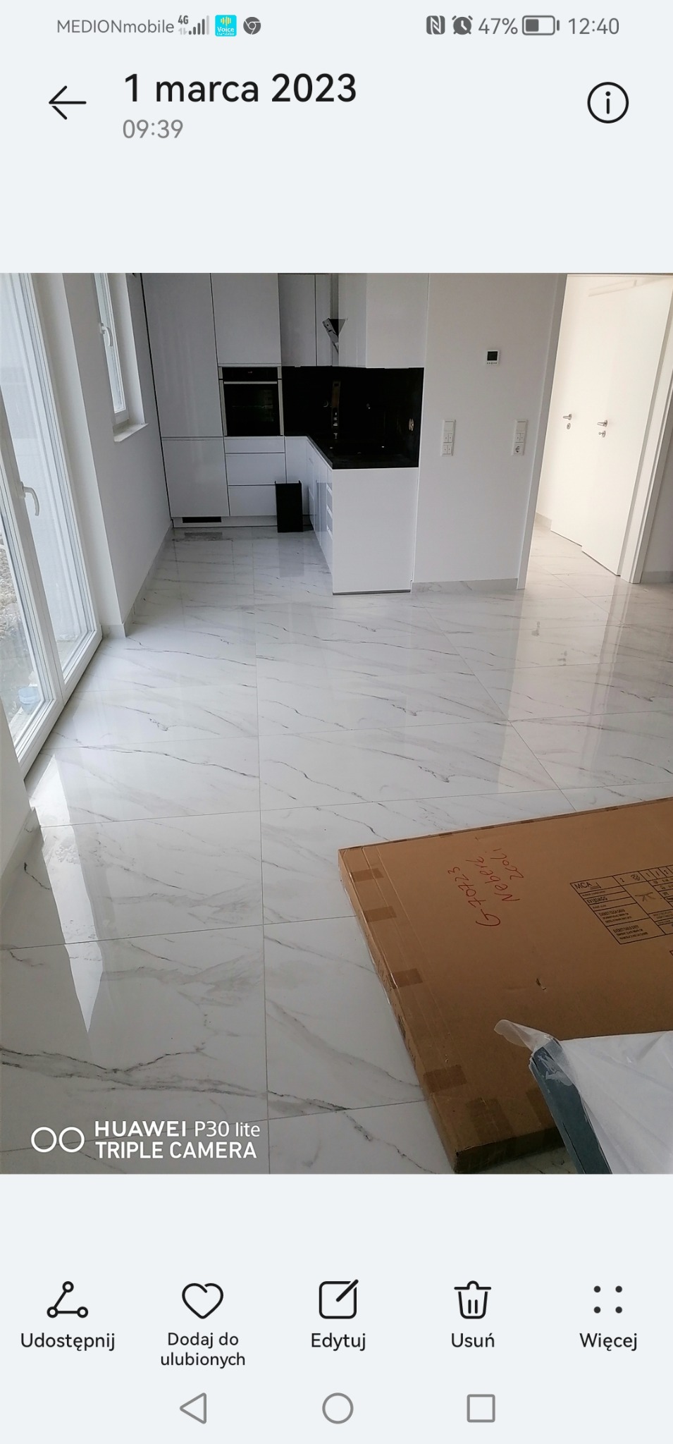 Błyszcząca, biała marmurowa podłoga w nowym, minimalistycznym wnętrzu z białą zabudową kuchenną i kartonem z napisem 'G-70723 Neberg Zoli' leżącym na podłodze.