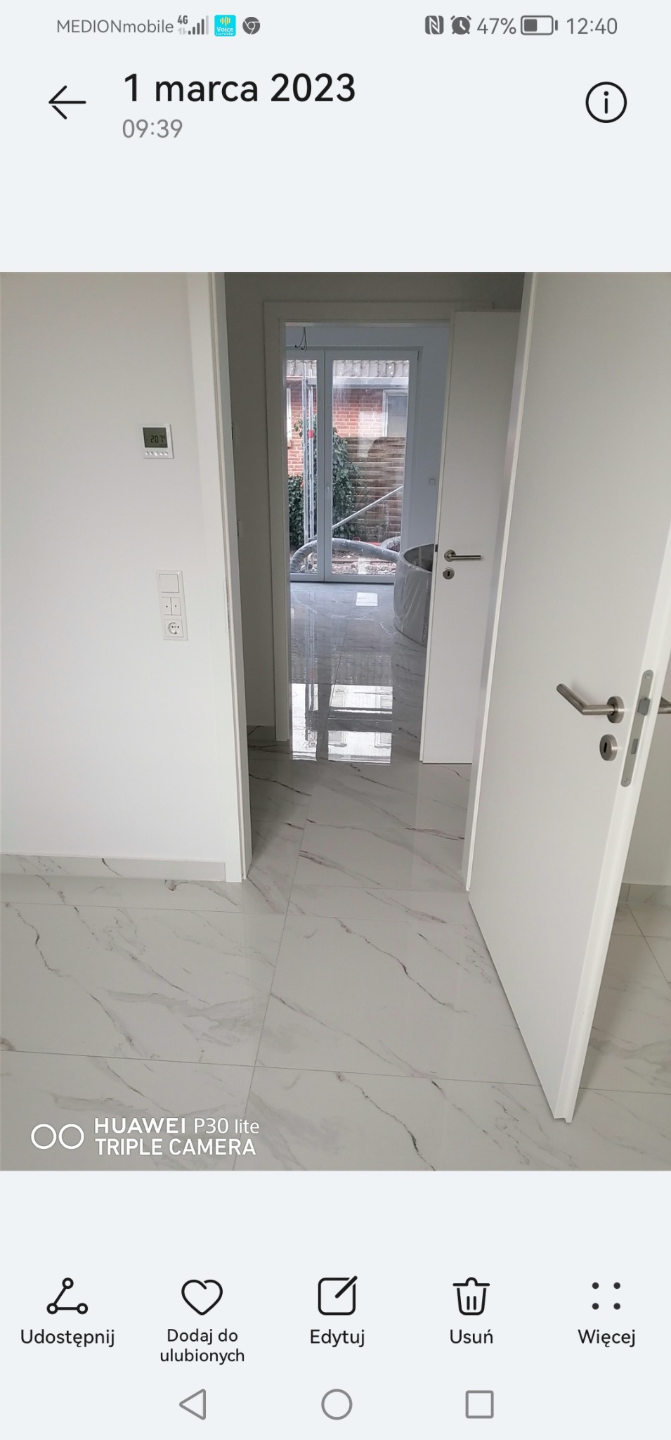 Białe, błyszczące płytki imitujące marmur na podłodze w pustym pomieszczeniu, widok na otwarte drzwi balkonowe i fragment wanny zabezpieczonej folią.