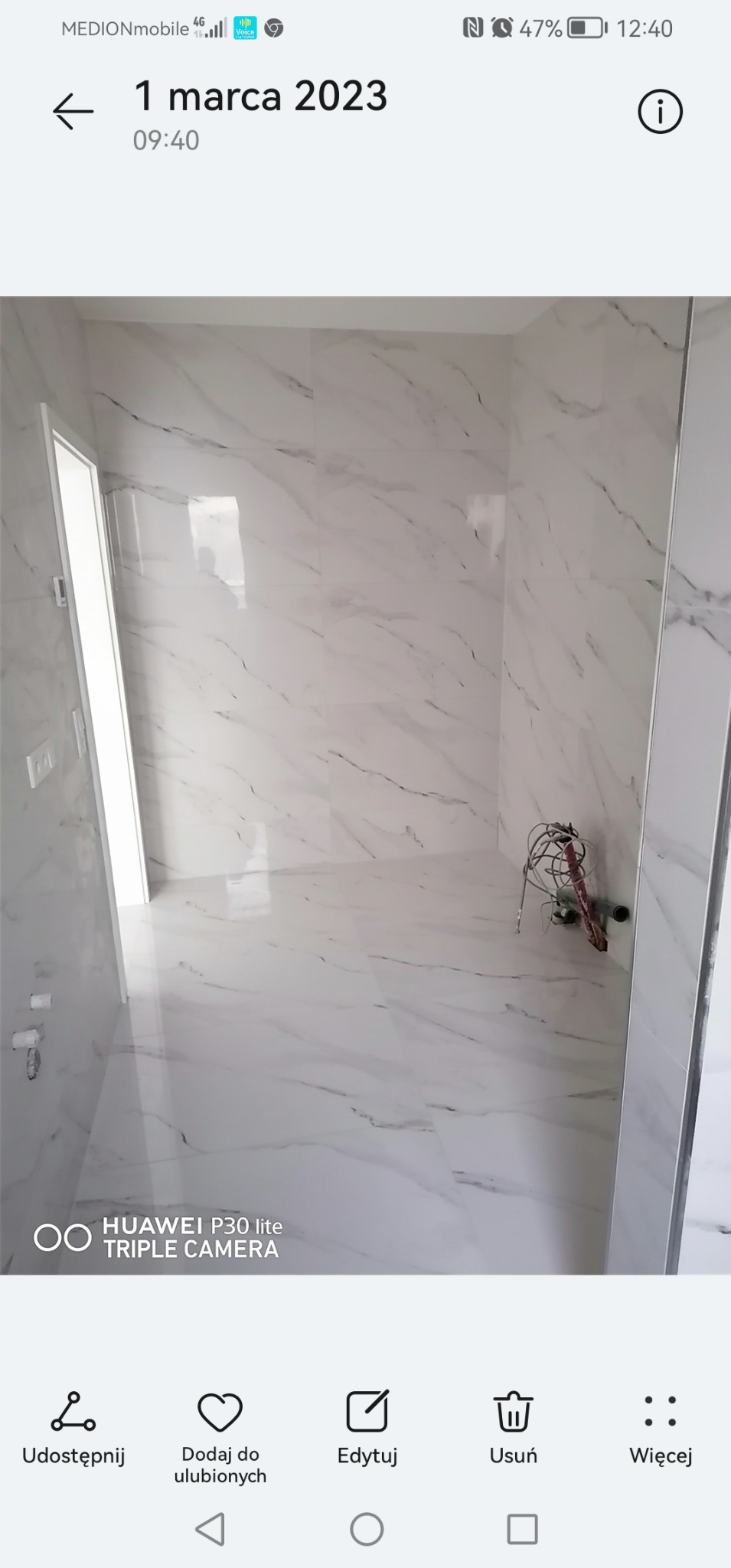 Wnętrze łazienki w trakcie remontu, widoczne białe płytki imitujące marmur na ścianach i podłodze, wystające przewody instalacji elektrycznej i hydraulicznej, niedokończone wykończenie.