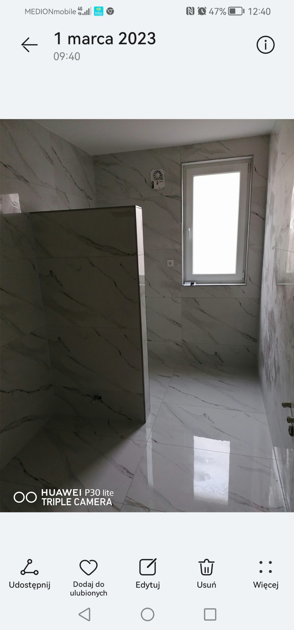 Nowoczesna łazienka z białymi płytkami imitującymi marmur, kabina prysznicowa typu walk-in z widocznymi elementami montażowymi, okno z białymi ramami, wentylator kanałowy na ścianie.