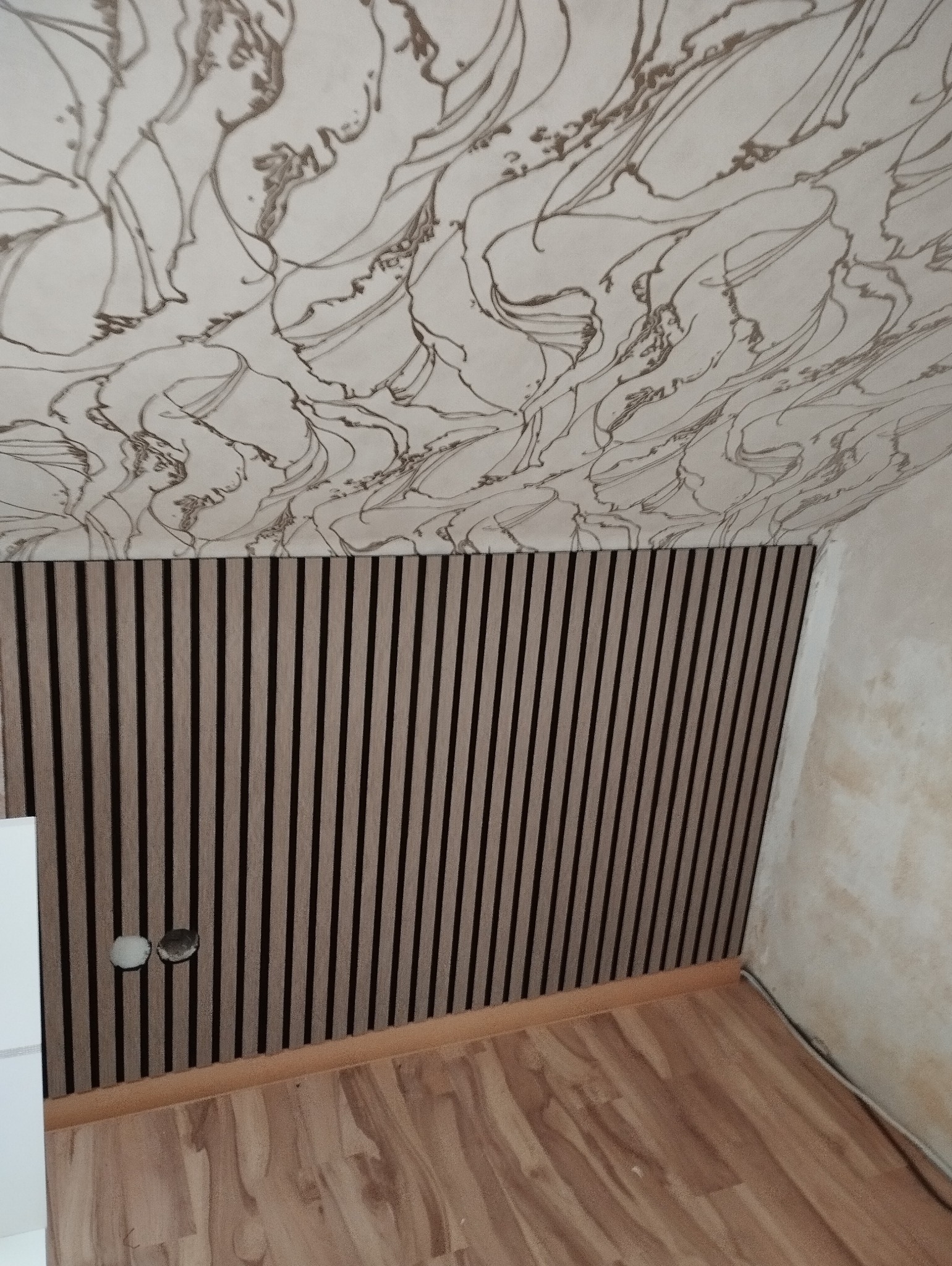 Panel ścienny z drewnianymi lamelami w brązowo-czarnych odcieniach, połączony z sufitem pokrytym wzorzystą tapetą w odcieniach beżu i brązu, widoczne gniazdka elektryczne i fragment podłogi z paneli.
