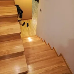 Widok z góry na jasne, dębowe schody z podstopnicami, prowadzące w dół, z widocznym fragmentem korytarza z obuwiem i odzieżą.