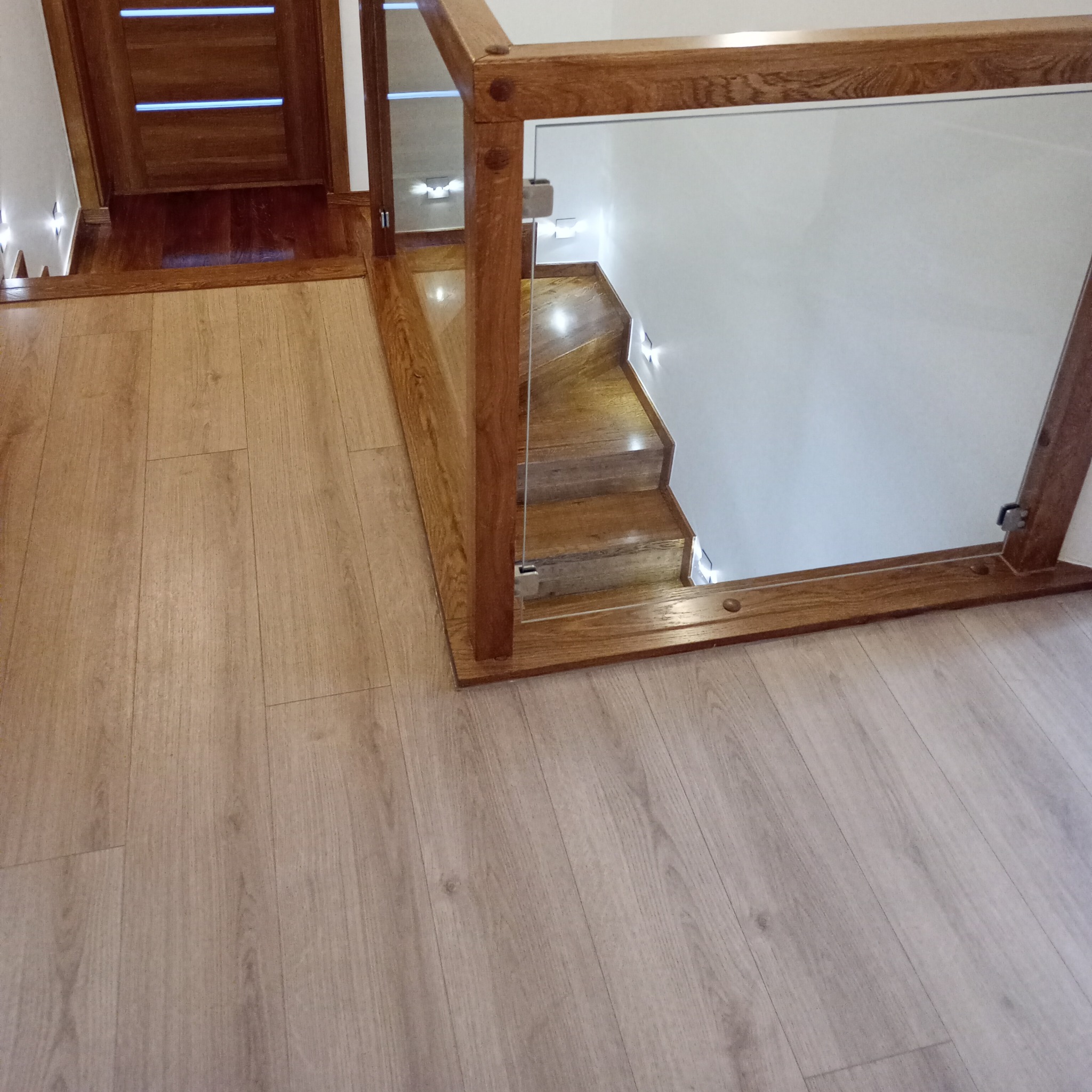 Wnętrze z drewnianymi schodami z oświetleniem punktowym i szklaną balustradą, widok z góry na podłogę z paneli.