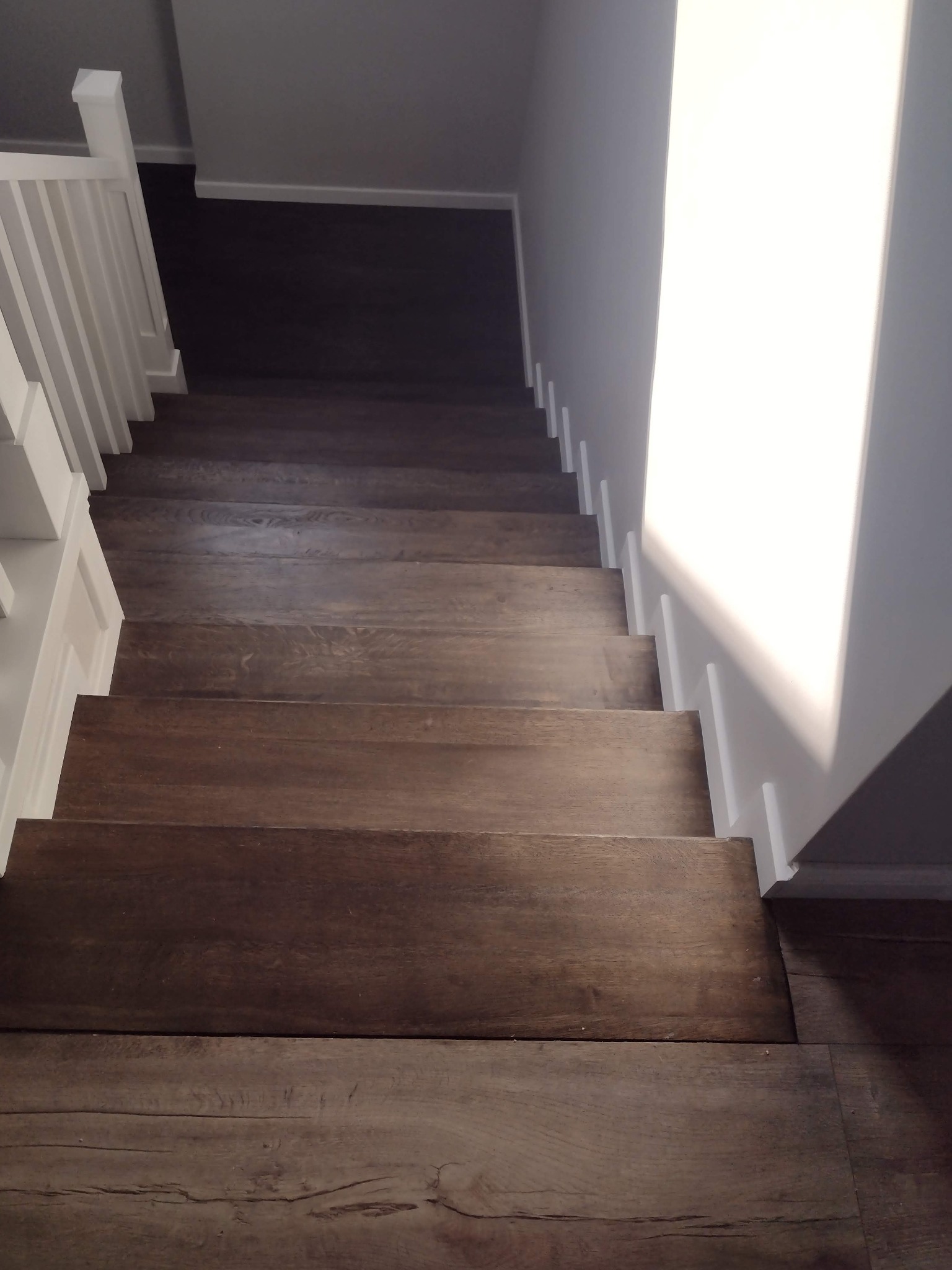 Widok z góry na brązowe stopnie z drewna, biała balustrada po lewej stronie, ściana z białymi listwami przypodłogowymi po prawej, oświetlone naturalnym światłem.