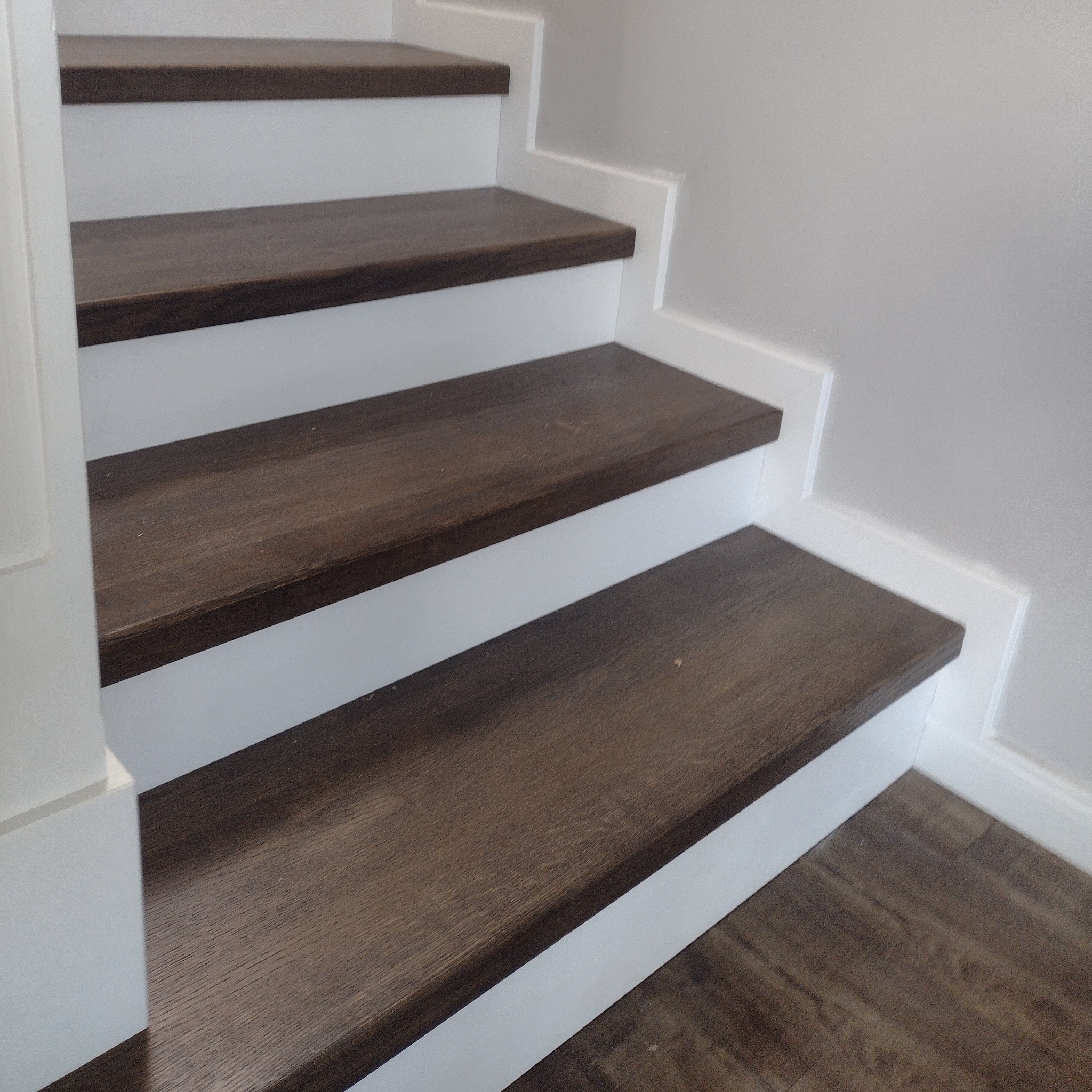 Ciemnobrązowe stopnie z drewna na białych podstopnicach, widok z dołu na fragment biegu schodów przy białej ścianie.