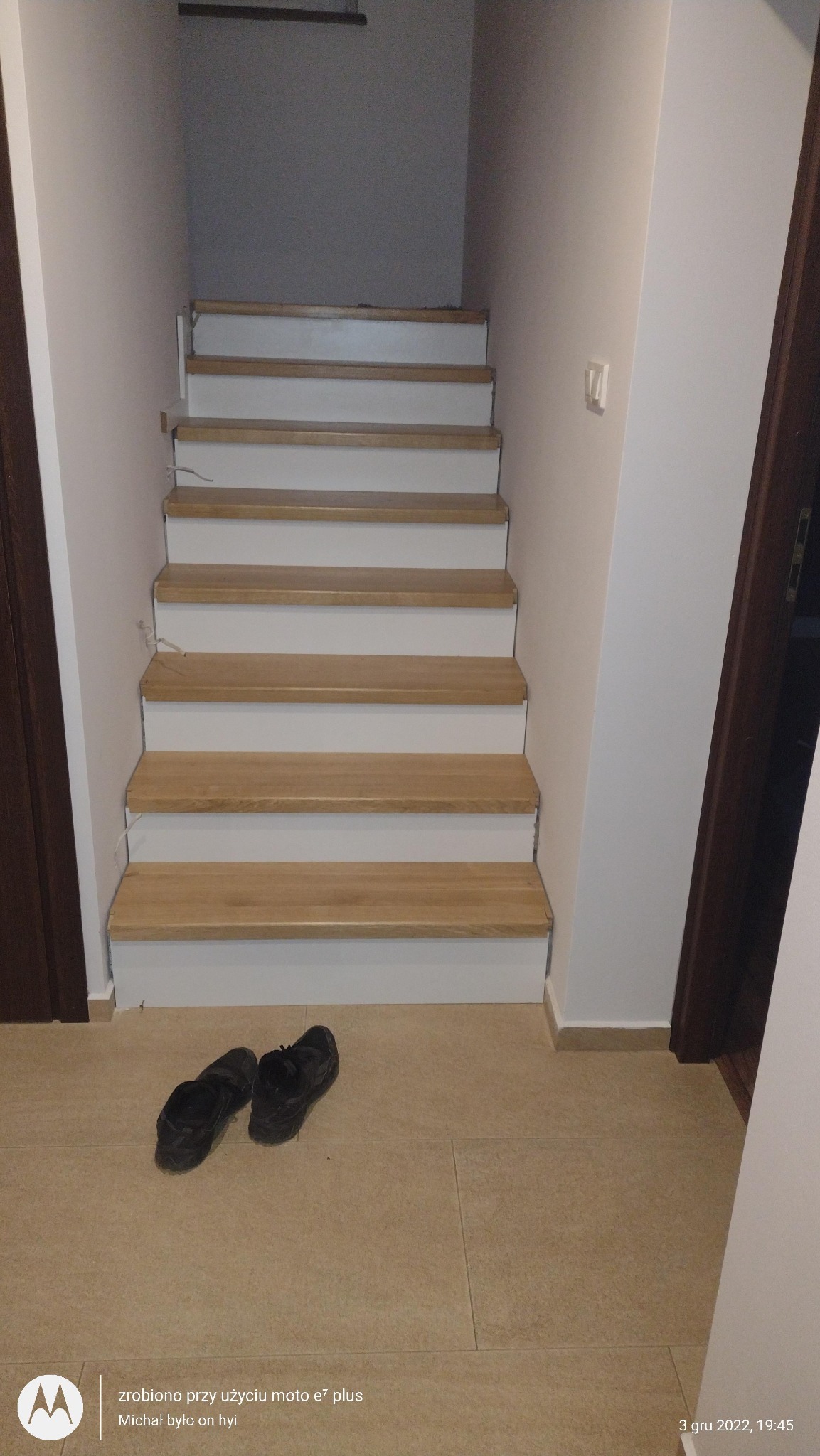 Białe schody z drewnianymi stopniami prowadzą w górę; na podłodze leżą czarne buty.