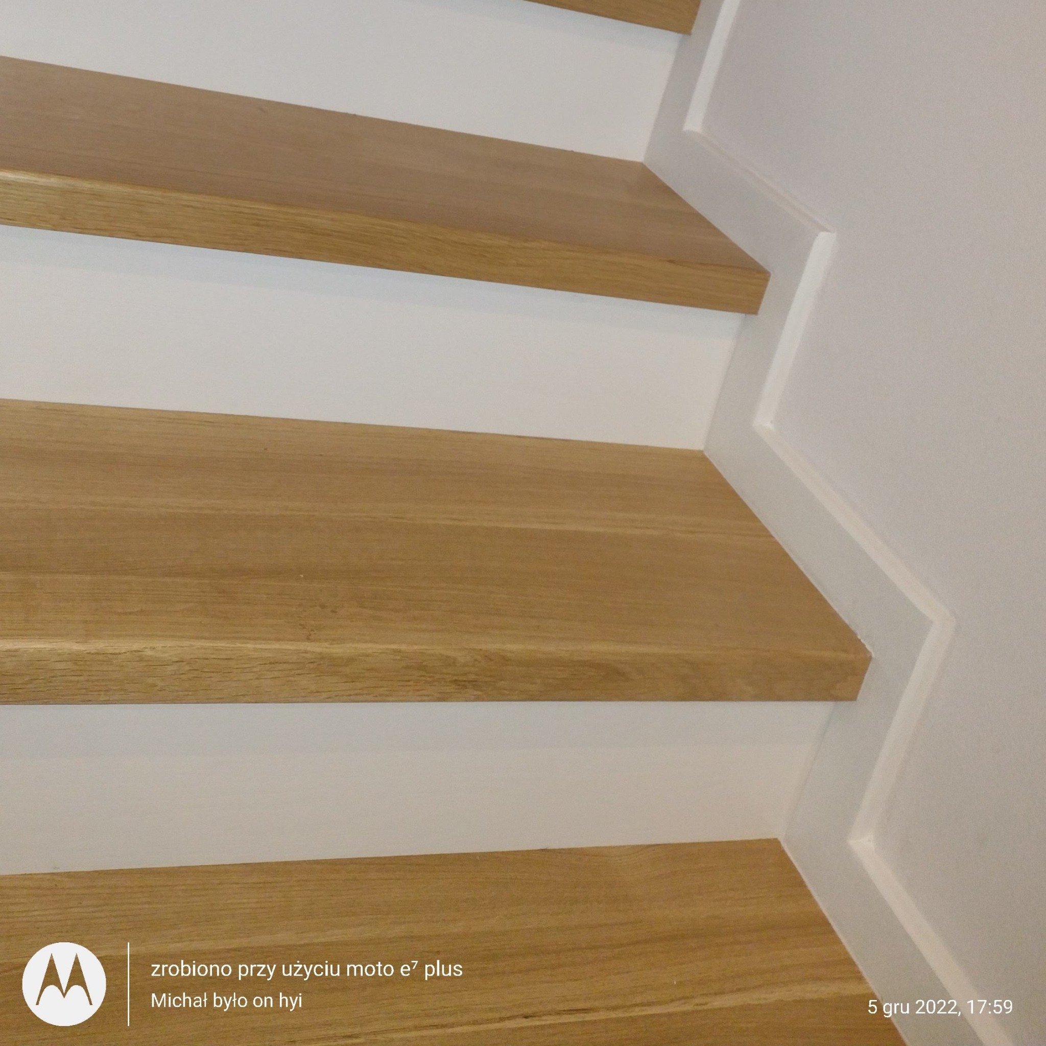 Fragment drewnianych schodów z białymi podstopnicami i listwami przypodłogowymi, widoczne trzy stopnie z bliska.