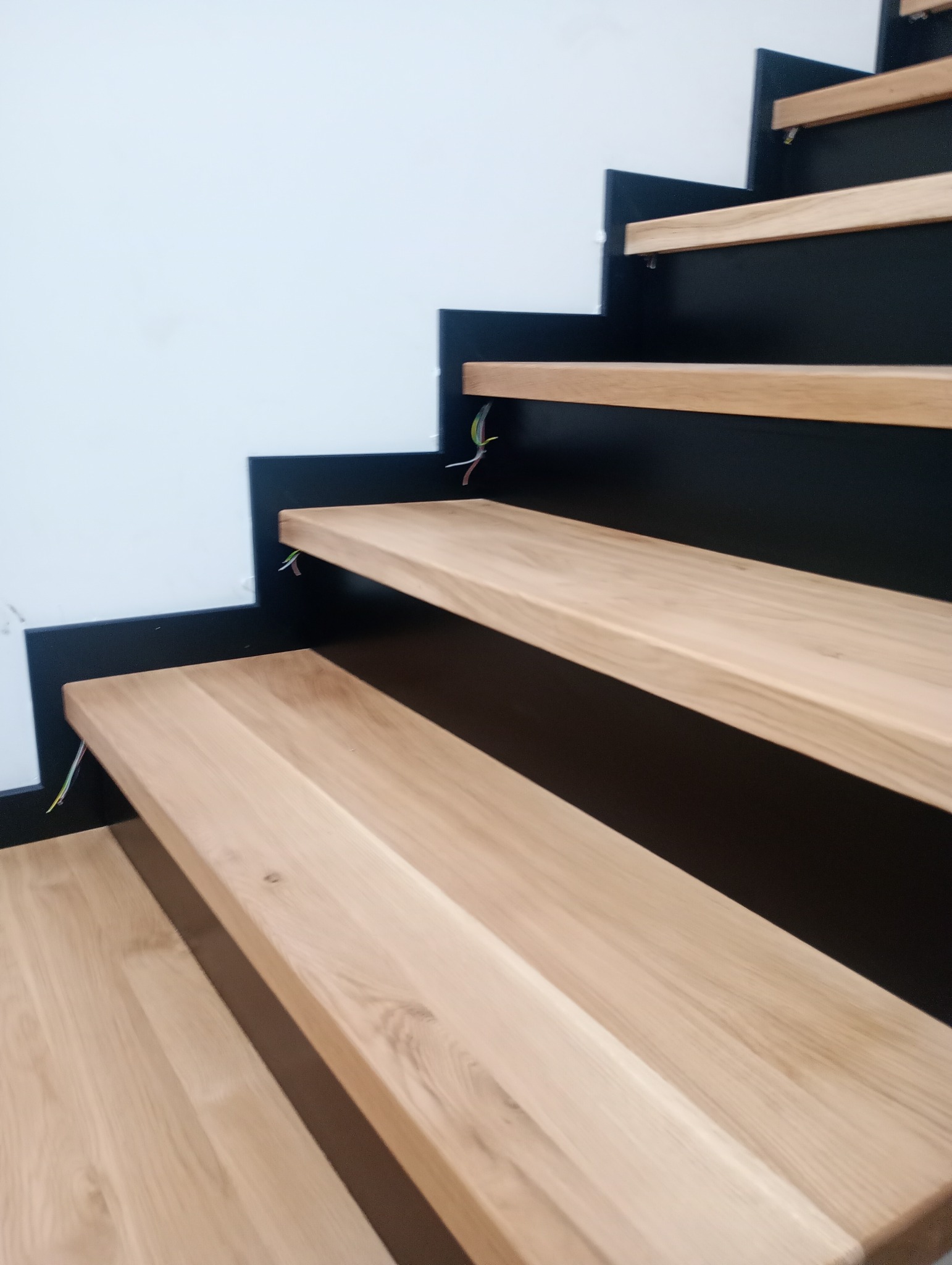 Surowe, jasne stopnie zamontowane na czarnej konstrukcji przy białej ścianie, widoczne kable elektryczne, realizacja z Węgierskiej Górki w trakcie montażu.