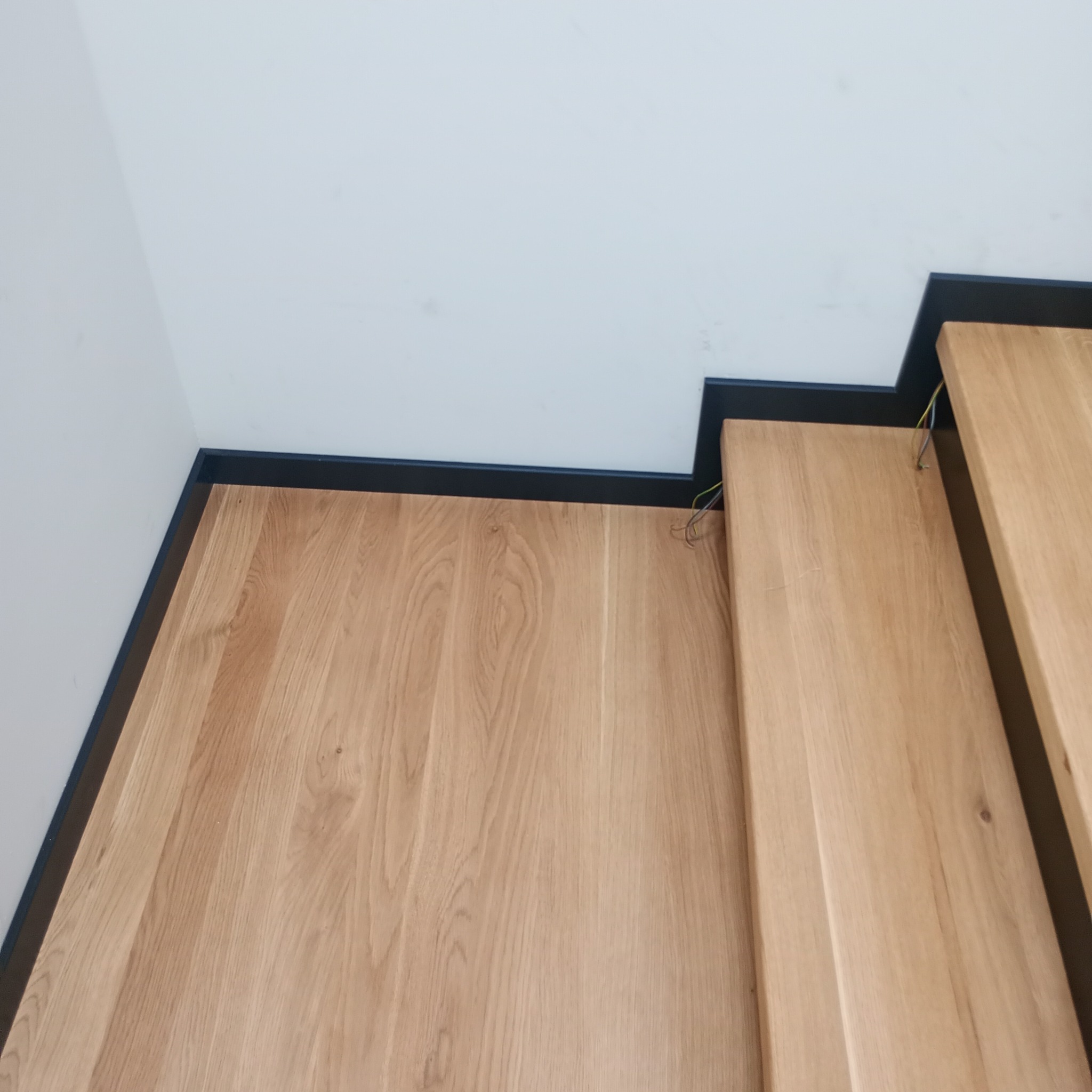 Fragment drewnianych schodów z czarną listwą przypodłogową, widoczne wystające kable elektryczne.