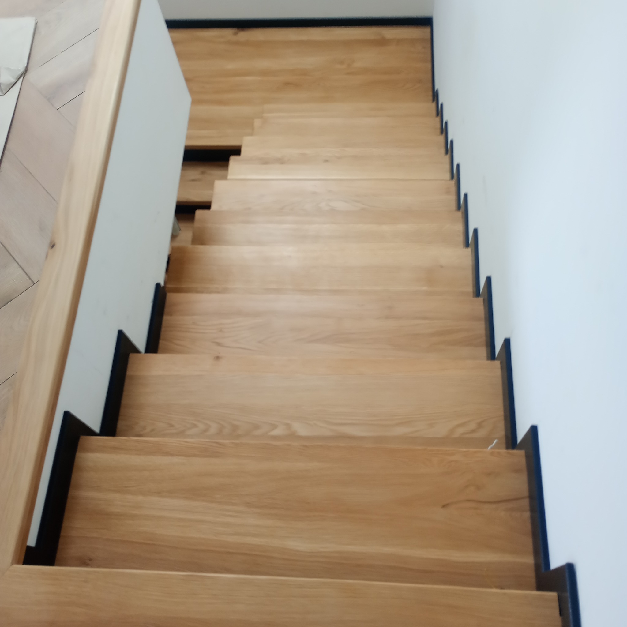 Widok z góry na jasne, dębowe stopnie schodów z czarnymi podstopnicami, z białymi ścianami po bokach i drewnianą podłogą na górnym poziomie.