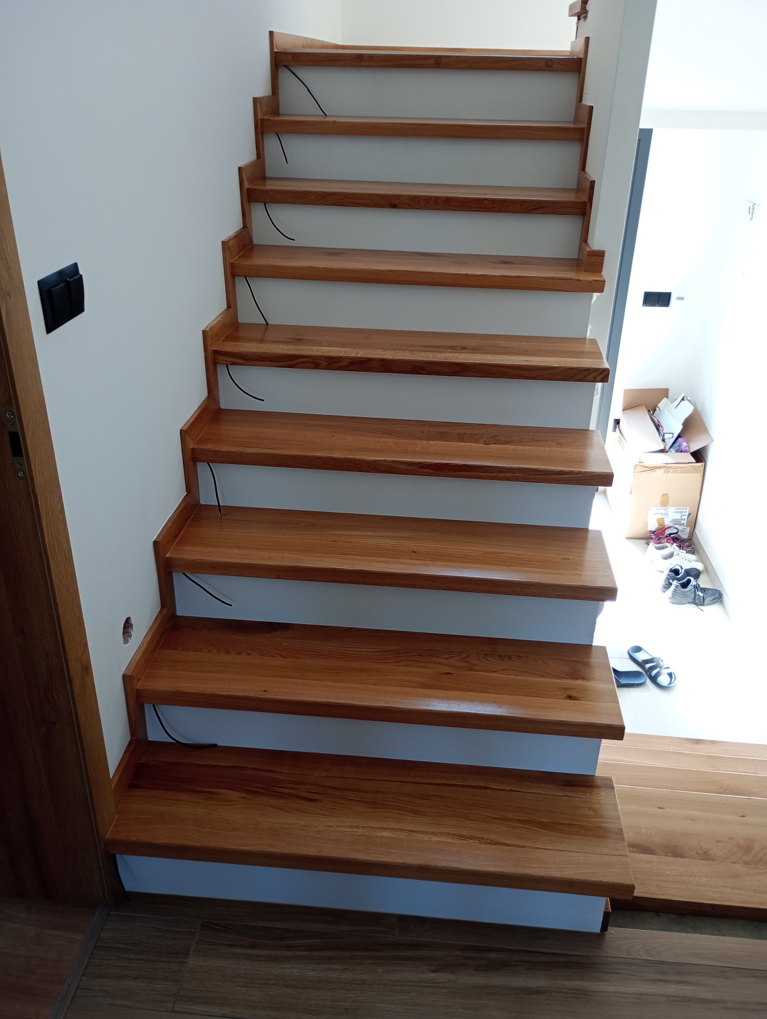 Nowoczesne schody z drewna dębowego z białymi podstopnicami, widoczne przewody elektryczne do oświetlenia, w tle widoczne kartony i obuwie.