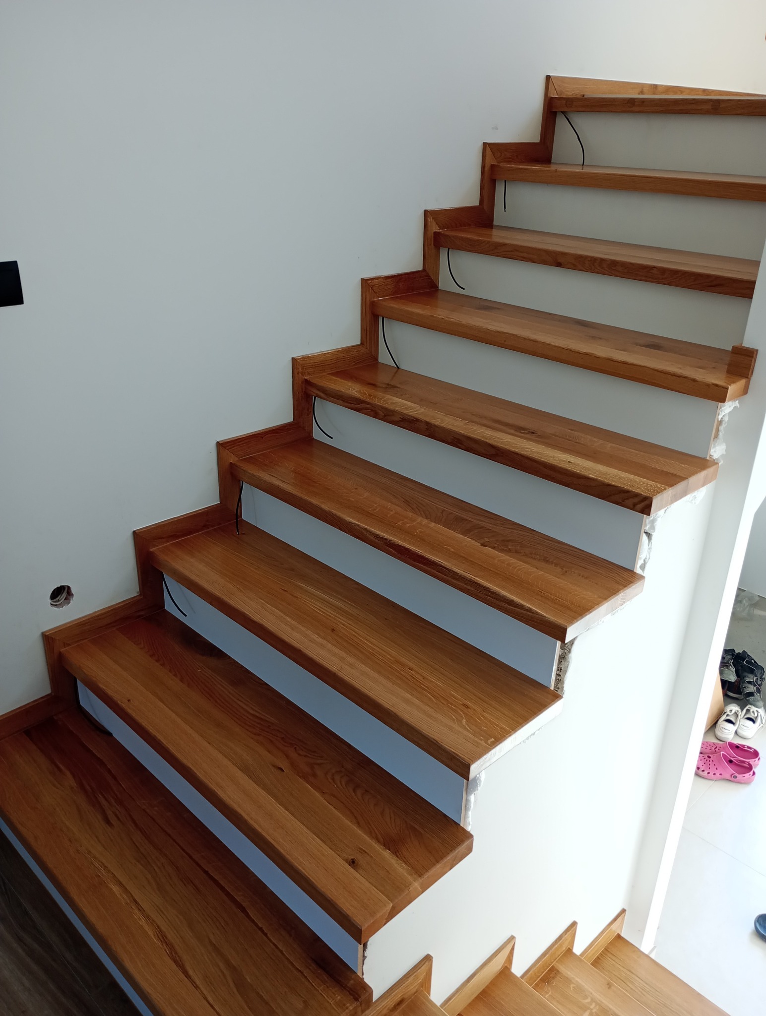 Nowoczesne, dębowe schody z widocznymi niedociągnięciami wykończeniowymi przy ścianie i wystającymi kablami spod stopni w trakcie montażu.