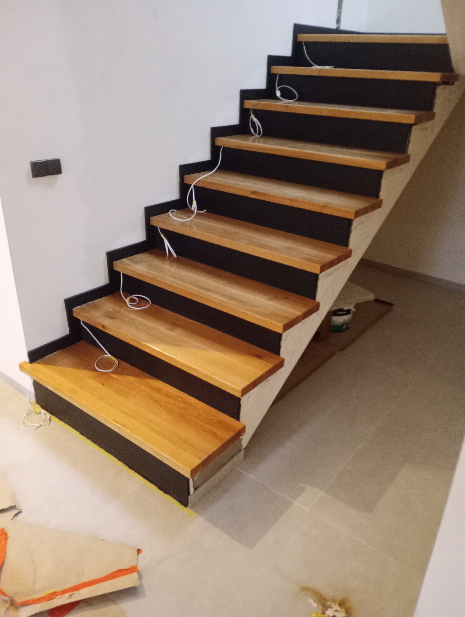 Stopnie schodów z jasnego drewna z czarnymi podstopnicami, widoczne kable elektryczne, podłoga w trakcie wykańczania.