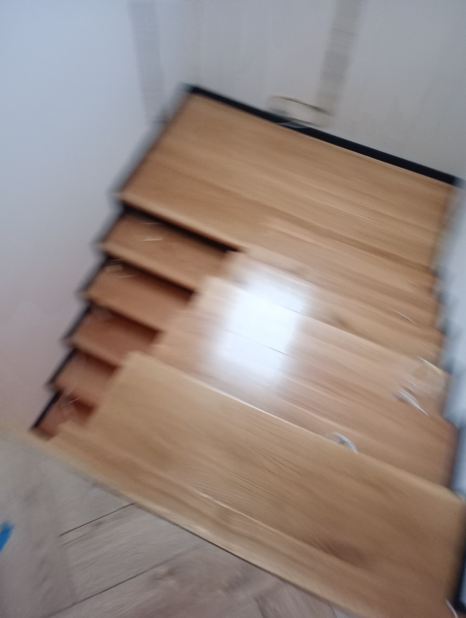 Rozmyte ujęcie z góry na drewniane schody z czarnymi krawędziami, widoczne białe ściany i fragment podłogi na dole kadru.