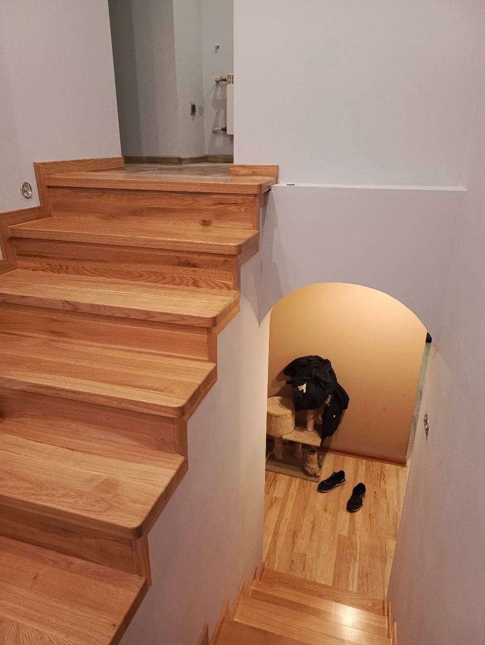 Jasne, drewniane schody z zaokrąglonymi krawędziami stopni, prowadzące do pomieszczenia z niskim, łukowym wejściem, gdzie widoczny jest bałagan: ubrania na stojaku i buty na podłodze.