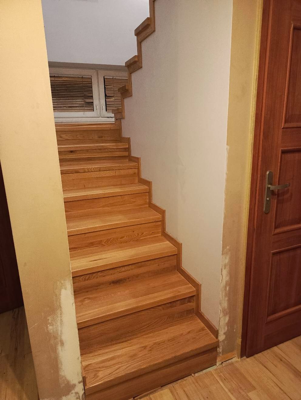Jasne, drewniane schody z widocznymi słojami drewna, prowadzące do góry, z oknem w tle i drzwiami po prawej stronie. Ściana obok schodów jest pomalowana na biało, a fragment ściany obok drzwi ma...
