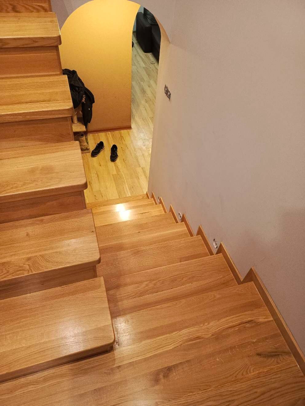 Widok z góry na jasne, dębowe schody z podstopnicami, prowadzące w dół, z widocznym fragmentem korytarza z obuwiem i odzieżą.