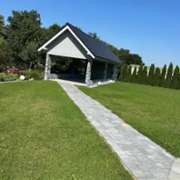 Murowana altana z ciemnym dachem, widok z perspektywy ścieżki z kostki brukowej prowadzącej przez trawnik w słoneczny dzień.