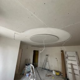 SolidConstruction - Sufit podwieszany z okrągłym elementem dekoracyjnym w trakcie montażu, widoczne szpachlowane łączenia płyt gipsowo-kartonowych. W tle drabina i worki z materiałami budowlanymi. Usługi w Gnieźnie.