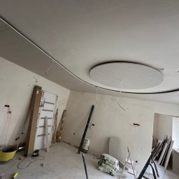 SolidConstruction - Wnętrze w trakcie remontu: widoczny sufit z dekoracyjną, okrągłą konstrukcją z płyt karton-gips, ściany przygotowane do wykończenia, worki z zaprawą i narzędzia budowlane na podłodze,...