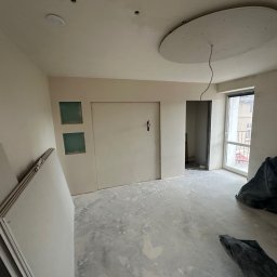 SolidConstruction - Wnętrze pokoju w trakcie remontu: ściany przygotowane do malowania, widoczne płyty gipsowo-kartonowe, podłoga z resztkami zaprawy, okrągła konstrukcja na suficie z wystającymi przewodami.