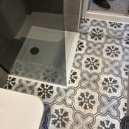 SolidConstruction - Wzorzysta, czarno-biała podłoga w łazience z kabiną prysznicową i fragmentem toalety; widoczne plamy farby na spodniach osoby stojącej w drzwiach.