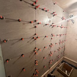 SolidConstruction - Ściana z szarych płytek podczas montażu, z użyciem pomarańczowych klinów i czarnych systemów poziomowania, widoczne niedoskonałości ścian i podłogi w trakcie remontu łazienki.