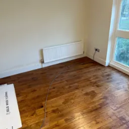Ukończona podłoga z drewna w pokoju z grzejnikiem, widoczne kartony z napisem 'Solid Wood Flooring' i przewód elektryczny na podłodze.