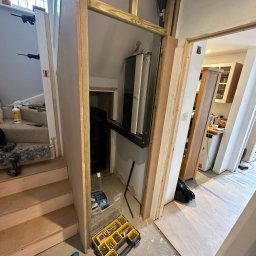 SolidConstruction - Adaptacja przestrzeni pod schodami: konstrukcja szkieletowa z drewna, narzędzia i materiały budowlane na podłodze, widok na korytarz i kuchnię w tle.