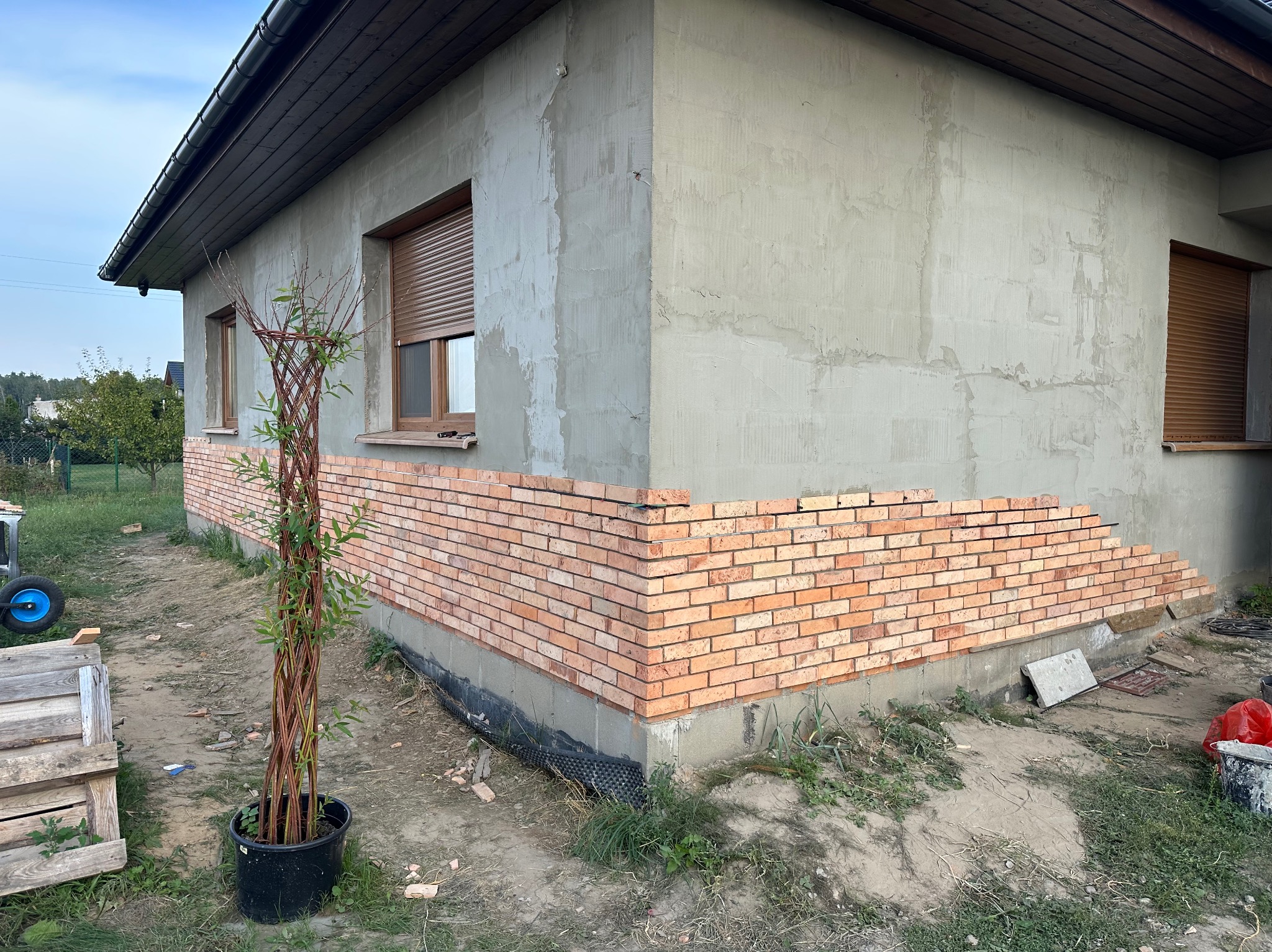 Fragment elewacji budynku w trakcie obkładania cegłą klinkierową, widoczne niedokończone prace na rogu budynku, surowy tynk na górnej części ściany, brązowe rolety okienne.