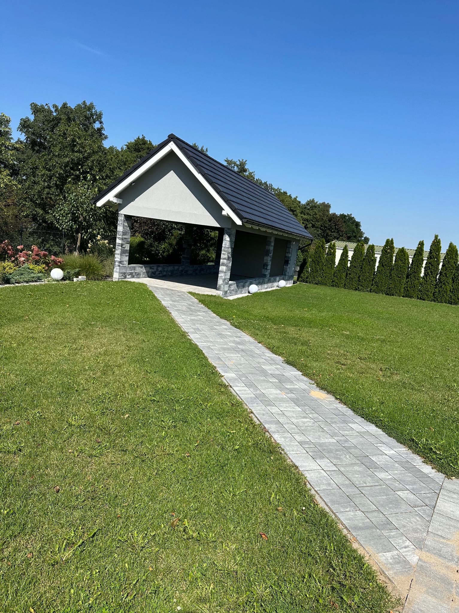 Murowana altana z ciemnym dachem, widok z perspektywy ścieżki z kostki brukowej prowadzącej przez trawnik w słoneczny dzień.