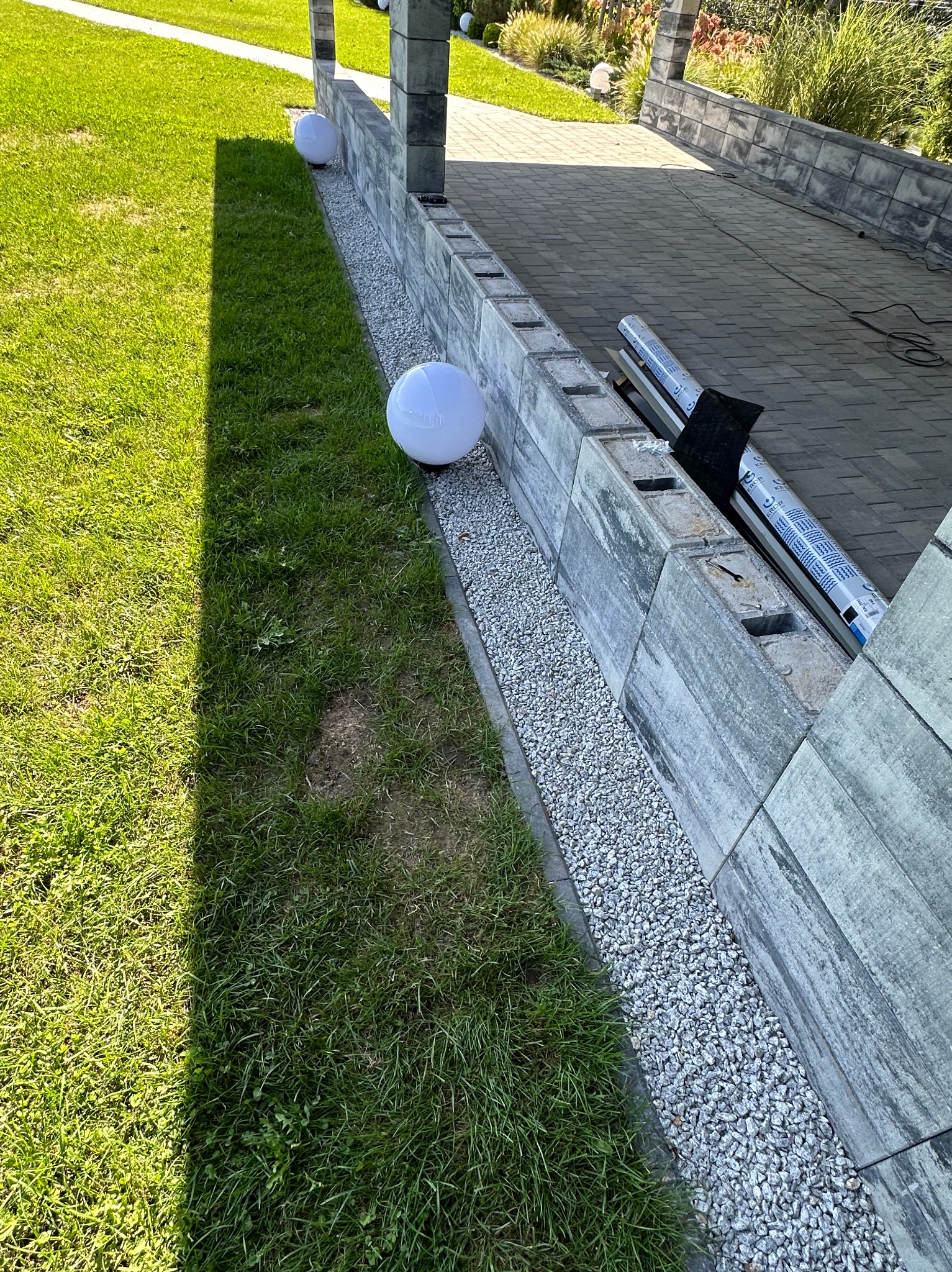 Fragment tarasu z kostki brukowej, obrzeże z szarego kamienia oddzielone od trawnika żwirem, dekoracyjne kule świetlne, w tle roślinność.