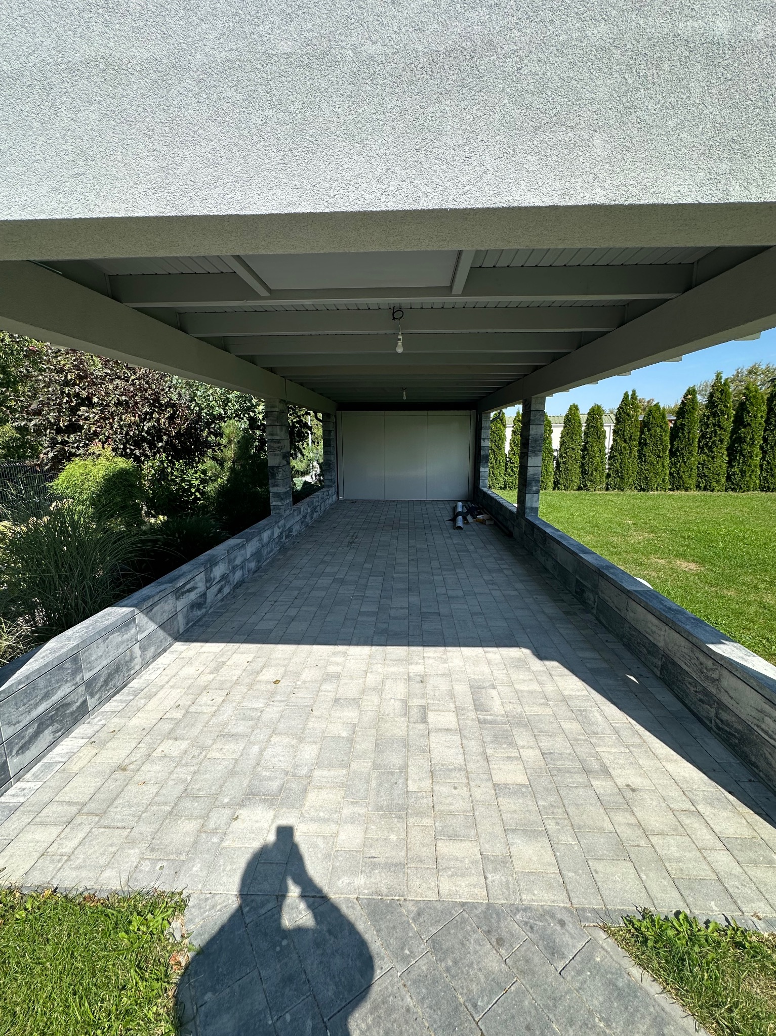 Zadaszone miejsce parkingowe z kostki brukowej, wykończone kamiennymi elementami, z widocznymi śladami świeżo zakończonych prac i zarysem osoby fotografującej w lewym dolnym rogu.