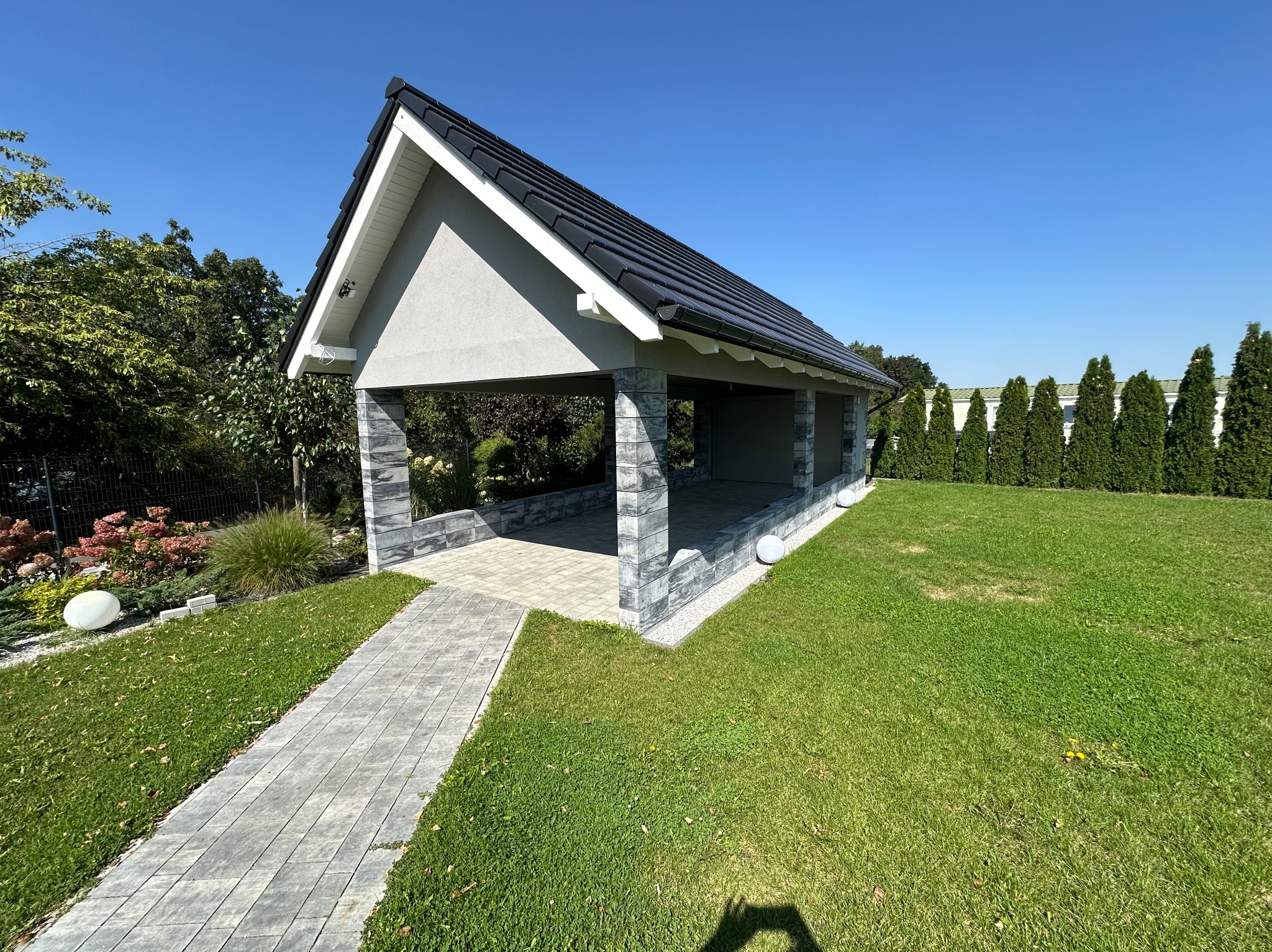 Nowoczesna altana z szarą elewacją i czarnym dachem, otoczona zielonym trawnikiem i tujami, z brukowaną ścieżką prowadzącą do wejścia.
