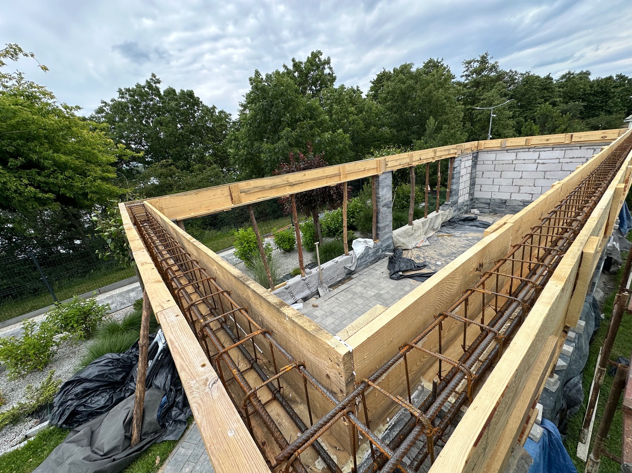 Budowa fundamentów pod budynek z drewnianym szalunkiem i zbrojeniem stalowym, widok z góry na plac budowy z drzewami w tle.