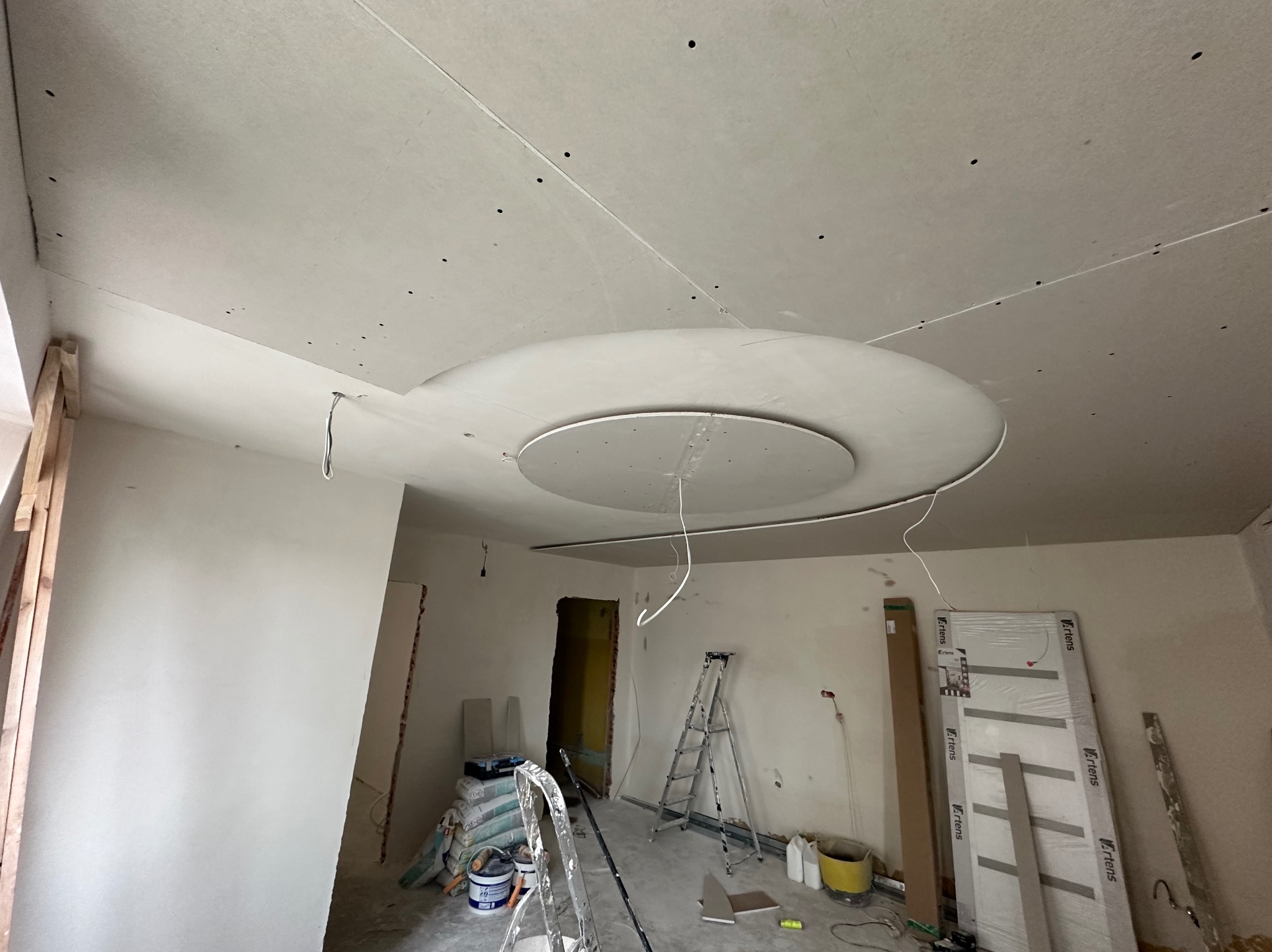 Sufit podwieszany z okrągłym elementem dekoracyjnym w trakcie montażu, widoczne szpachlowane łączenia płyt gipsowo-kartonowych. W tle drabina i worki z materiałami budowlanymi. Usługi w Gnieźnie.