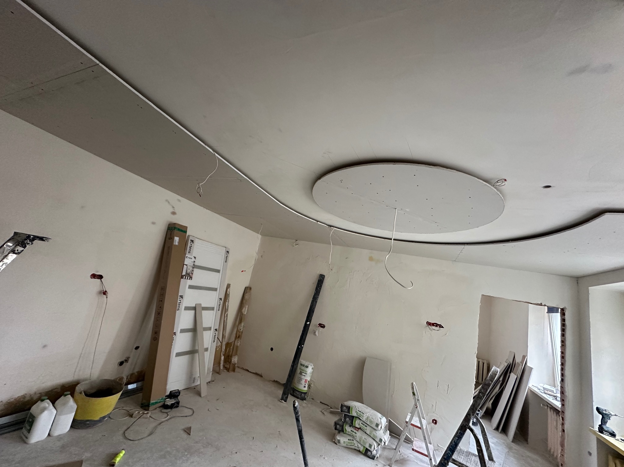 Wnętrze w trakcie remontu: widoczny sufit z dekoracyjną, okrągłą konstrukcją z płyt karton-gips, ściany przygotowane do wykończenia, worki z zaprawą i narzędzia budowlane na podłodze,...