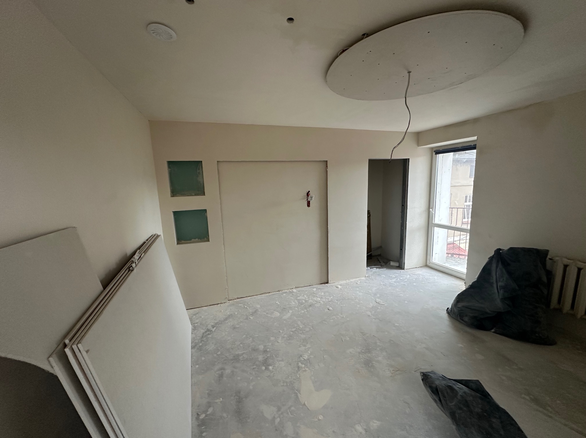 Wnętrze pokoju w trakcie remontu: ściany przygotowane do malowania, widoczne płyty gipsowo-kartonowe, podłoga z resztkami zaprawy, okrągła konstrukcja na suficie z wystającymi przewodami.