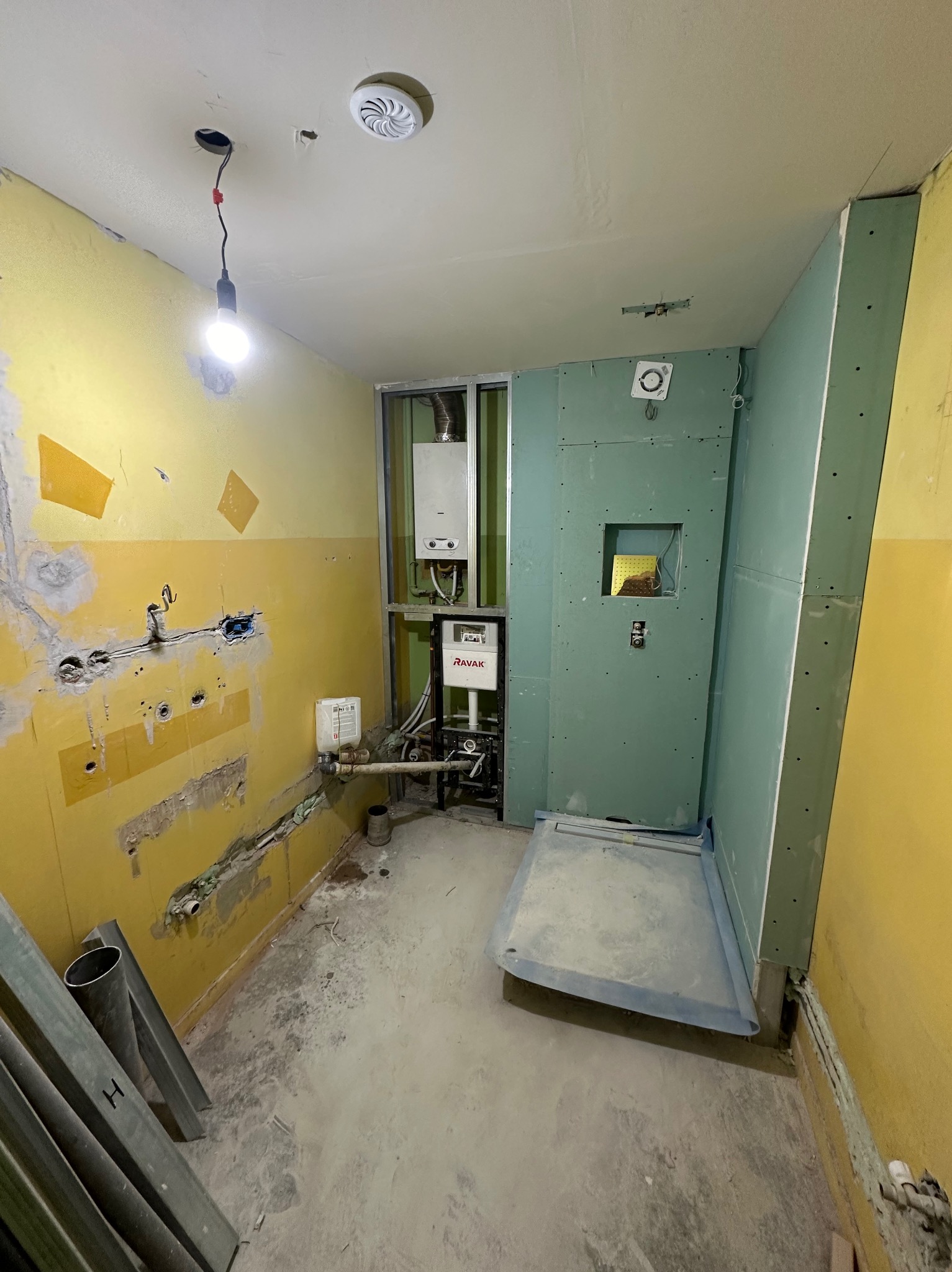 Niedokończona łazienka w trakcie remontu: żółte ściany z resztkami starej farby, instalacja grzewcza, prysznic z płytami gipsowo-kartonowymi, widoczne profile metalowe i wystające kable.