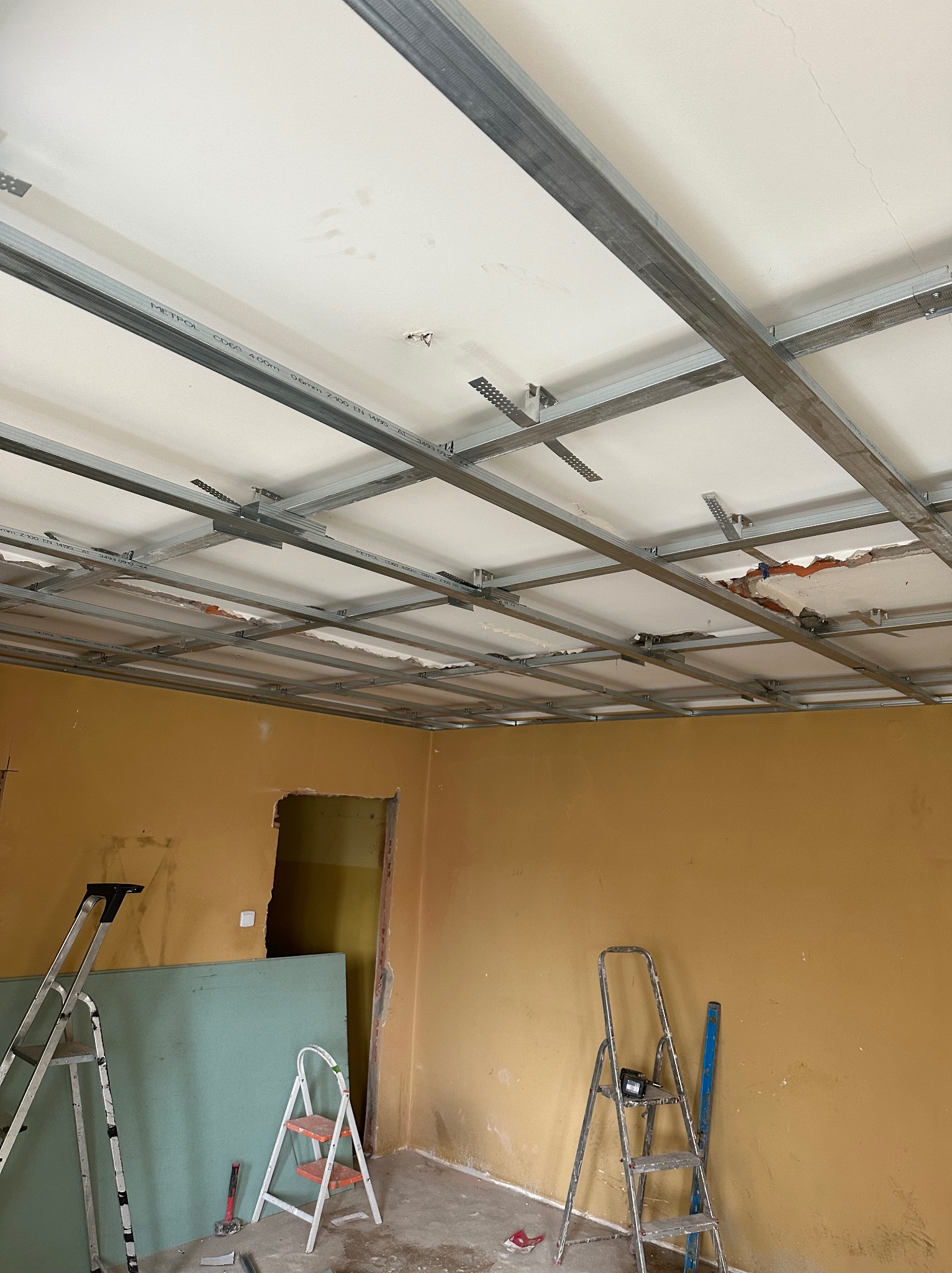 Montaż metalowej konstrukcji podwieszanego sufitu w pomieszczeniu z żółtymi ścianami, widoczne profile, płyty gipsowe i drabiny.