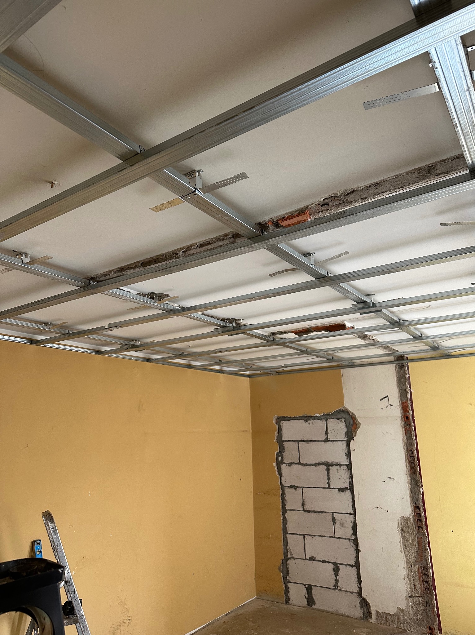 Sufit w trakcie montażu podwieszanego, widoczne metalowe profile konstrukcyjne, fragmenty starego sufitu i ściany w surowym stanie, żółte ściany, drabina oparta o ścianę.