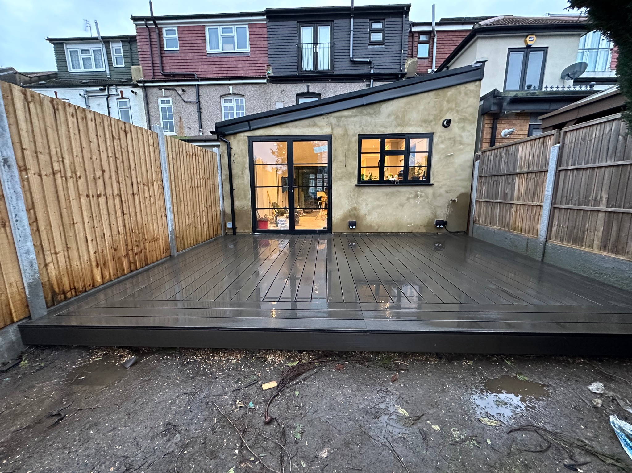 Nowo wybudowany, ciemnoszary taras kompozytowy przylegający do budynku z beżową elewacją. W tle widoczne zabudowania mieszkalne i drewniane ogrodzenie. Na pierwszym planie błotniste podłoże.
