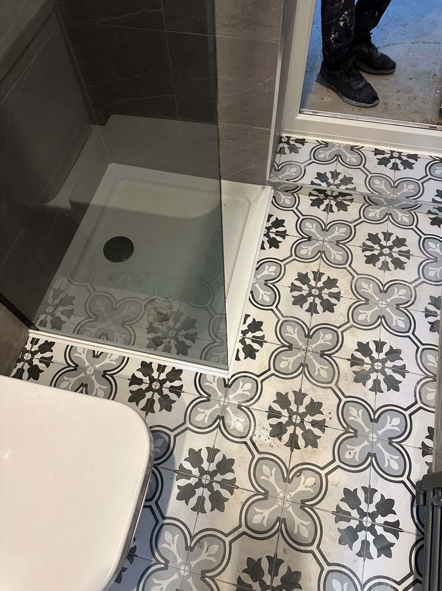 Wzorzysta, czarno-biała podłoga w łazience z kabiną prysznicową i fragmentem toalety; widoczne plamy farby na spodniach osoby stojącej w drzwiach.