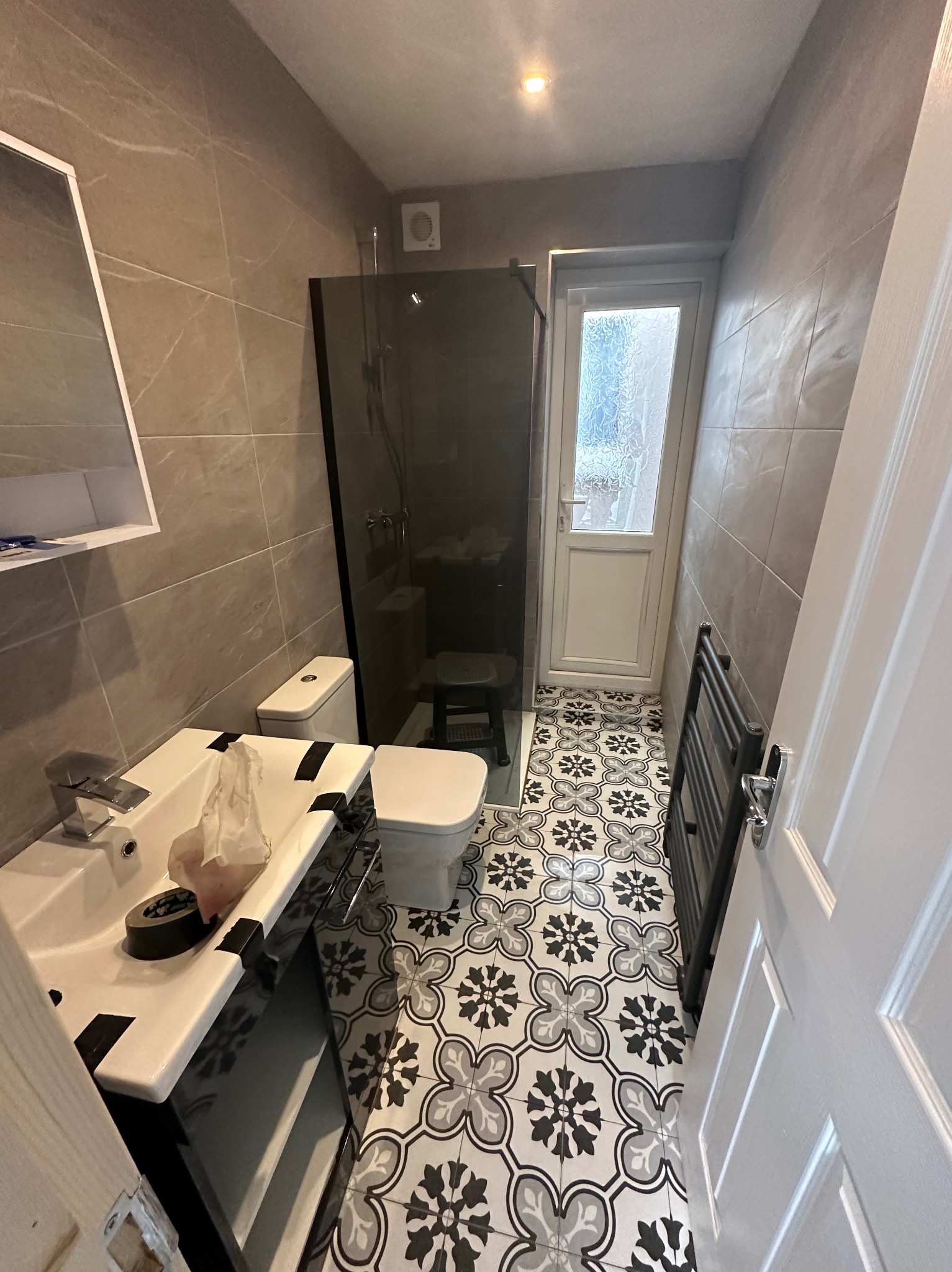 Wnętrze małej łazienki po remoncie: wzorzysta podłoga, kabina prysznicowa z czarnymi profilami, umywalka z szafką i toaleta w jasnych odcieniach szarości i bieli.