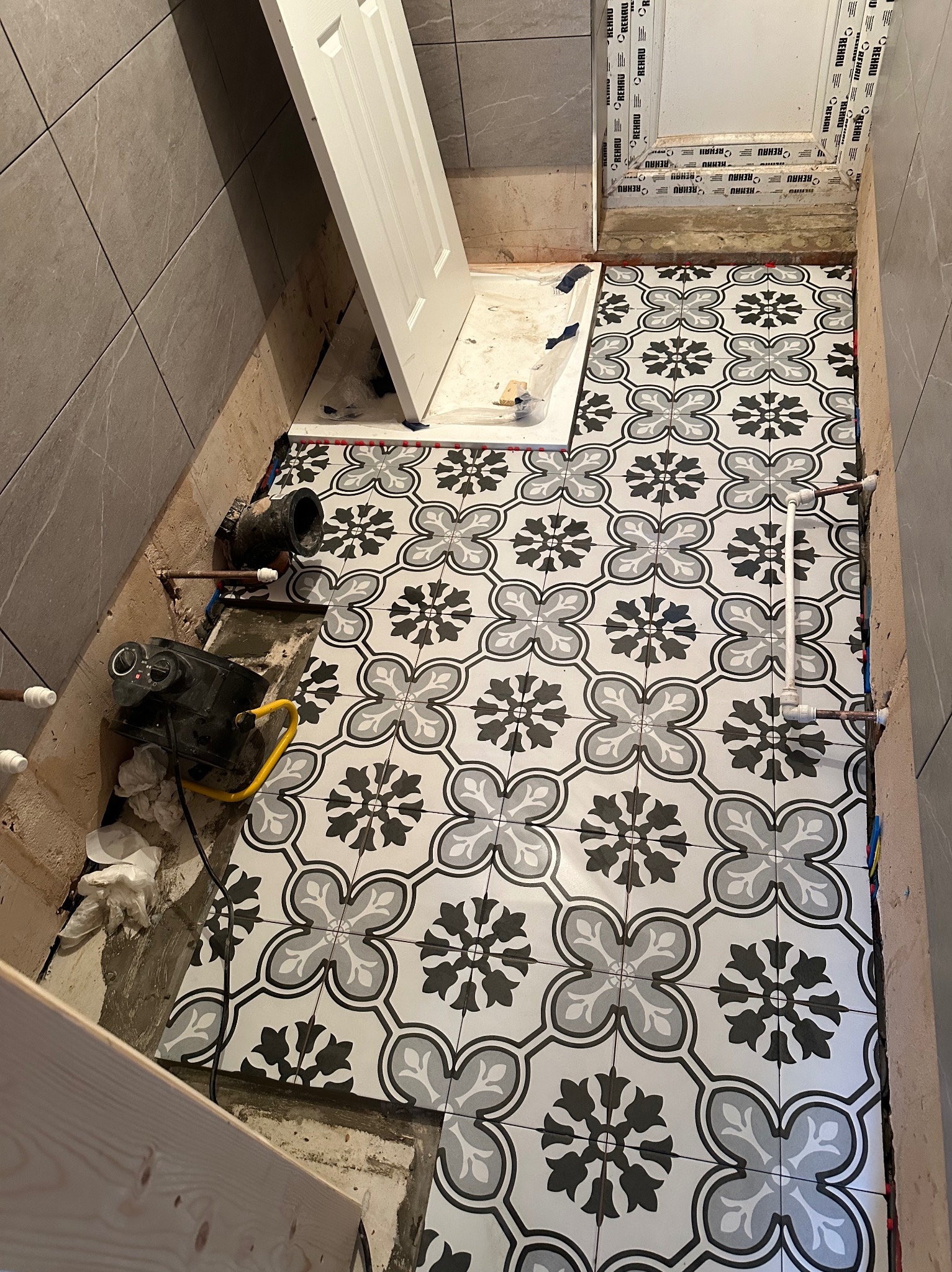 Proces układania wzorzystych, czarno-białych płytek podłogowych w łazience; widoczne narzędzia, rury i częściowo zamontowane drzwi.