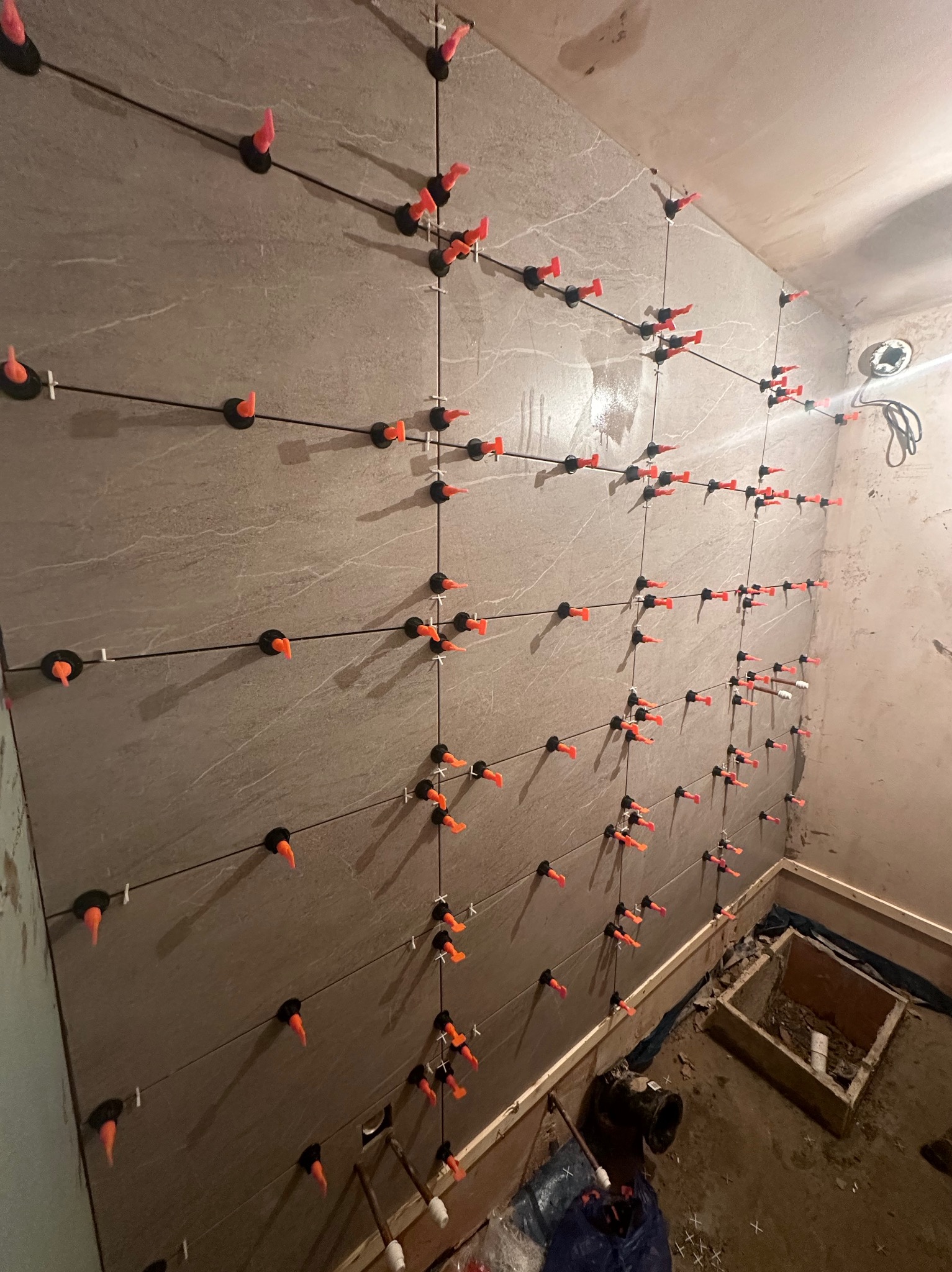 Ściana z szarych płytek podczas montażu, z użyciem pomarańczowych klinów i czarnych systemów poziomowania, widoczne niedoskonałości ścian i podłogi w trakcie remontu łazienki.