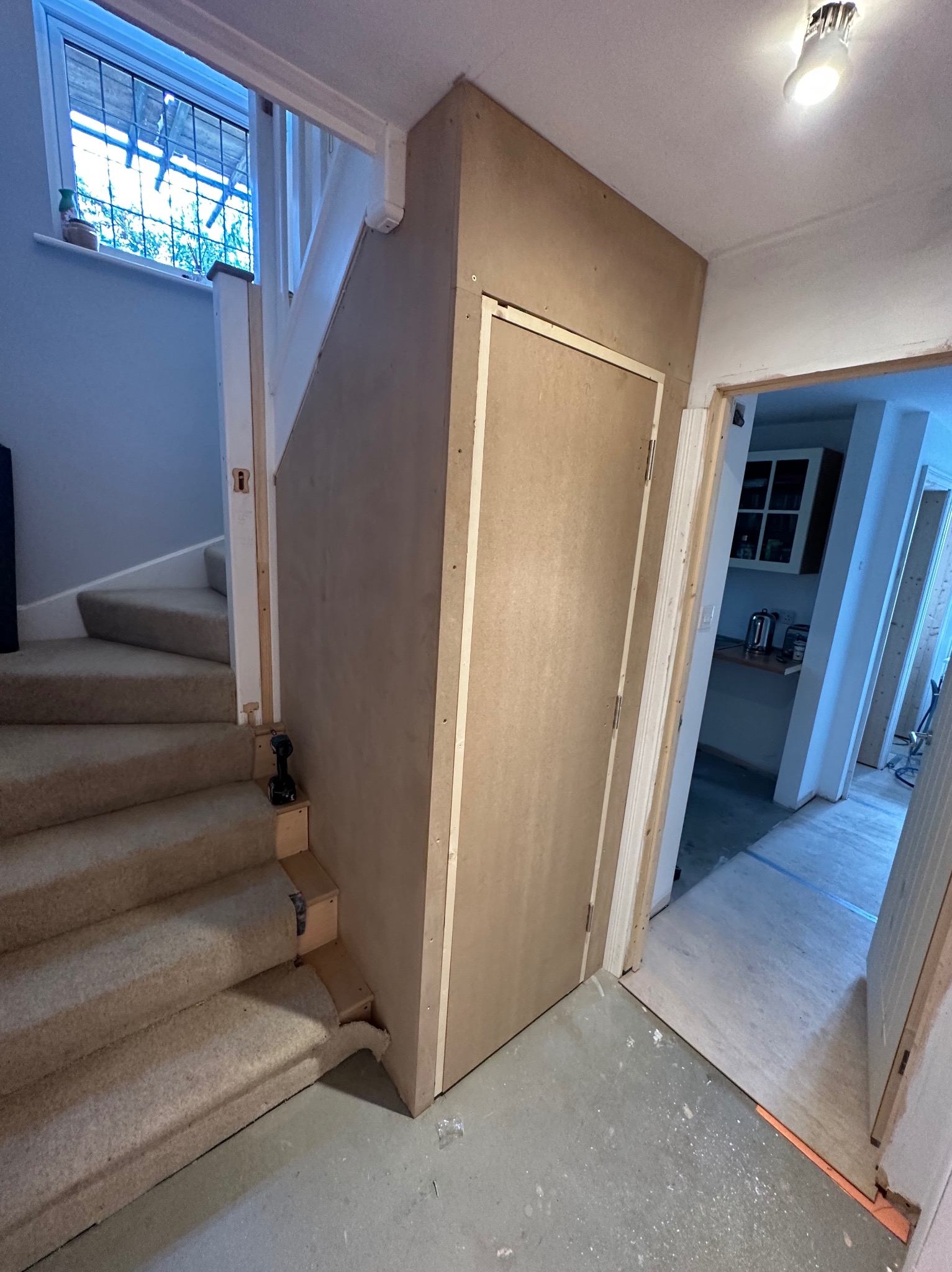 Zabudowa schodów z drzwiami wykonana z płyt MDF, widoczna wiertarka akumulatorowa na schodach, podłoga betonowa z drobnymi zabrudzeniami, widok na kuchnię w tle.