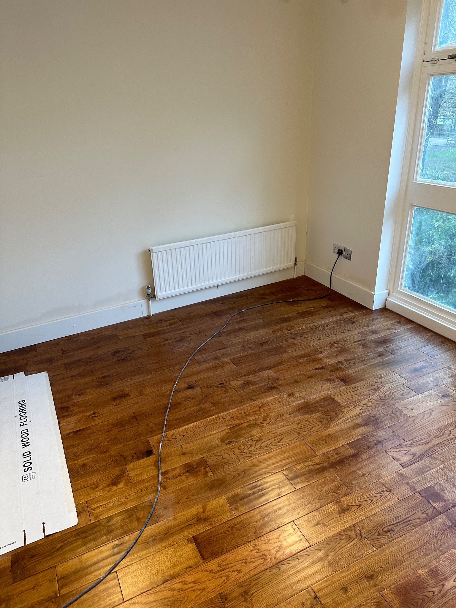 Ukończona podłoga z drewna w pokoju z grzejnikiem, widoczne kartony z napisem 'Solid Wood Flooring' i przewód elektryczny na podłodze.