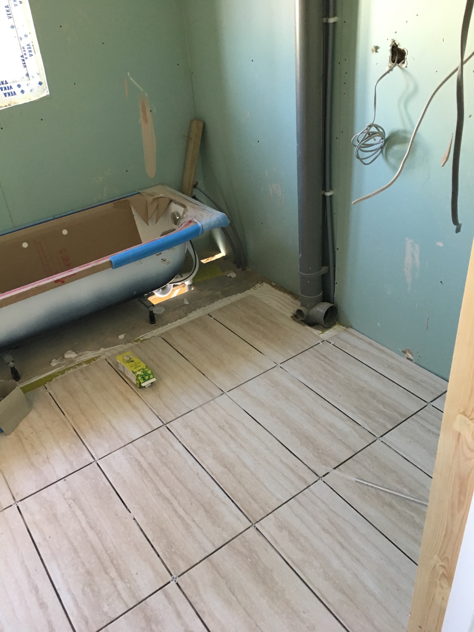 Wnętrze łazienki w trakcie remontu: ułożone beżowe płytki podłogowe z krzyżykami dystansowymi, przygotowana wanna z zabezpieczeniami, surowe ściany z instalacjami.